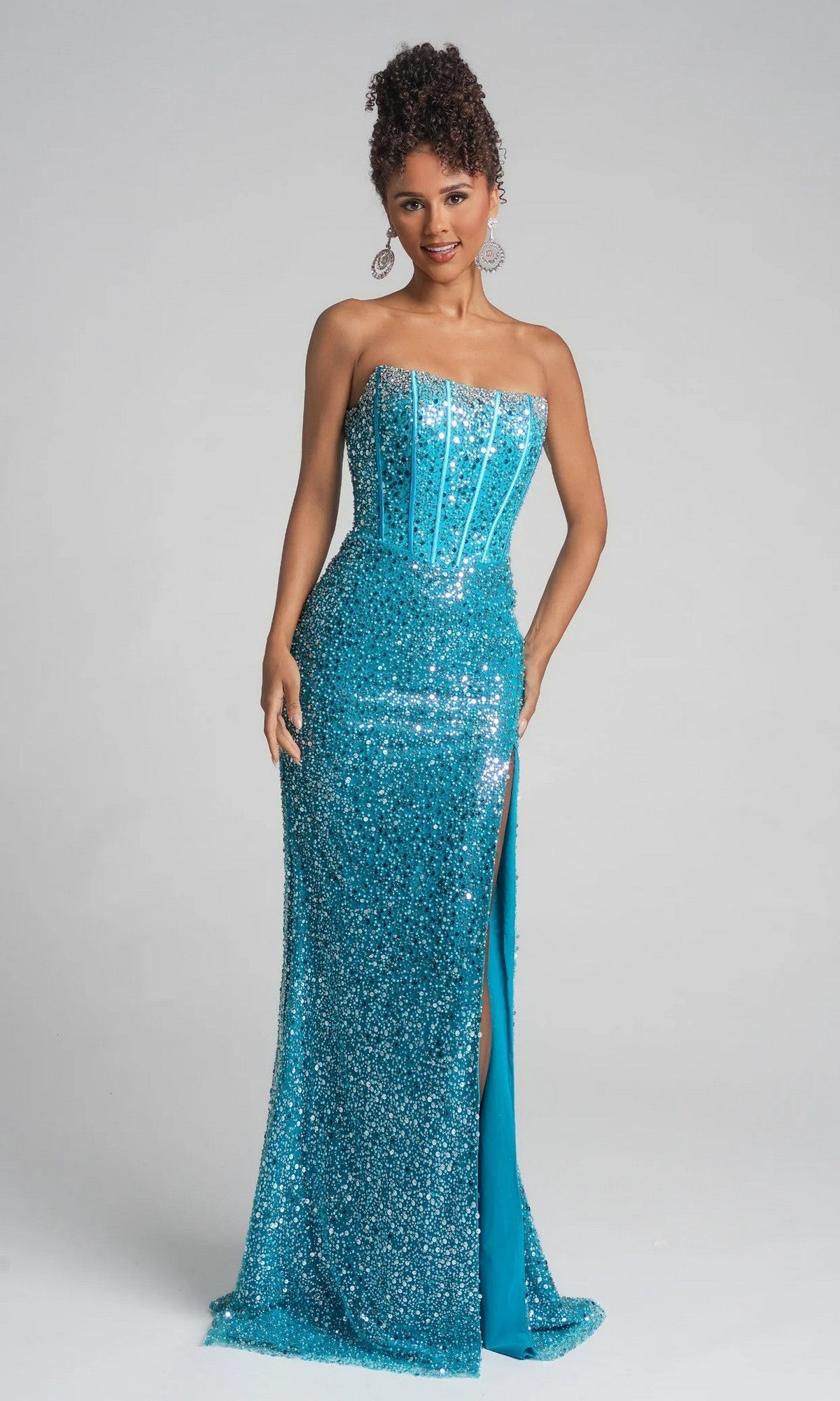 A woman stands against a plain gray background wearing the Portia and Scarlett XO054, a turquoise, floor-length formal dress featuring a strapless corset bodice, high slit, and sparkling sequins.